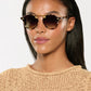 A woman with straight dark hair wears Krewe STL Nylon Sunglasses with flat nylon lenses and an oversized silhouette, paired with a beige knit top, facing forward against a plain light background.