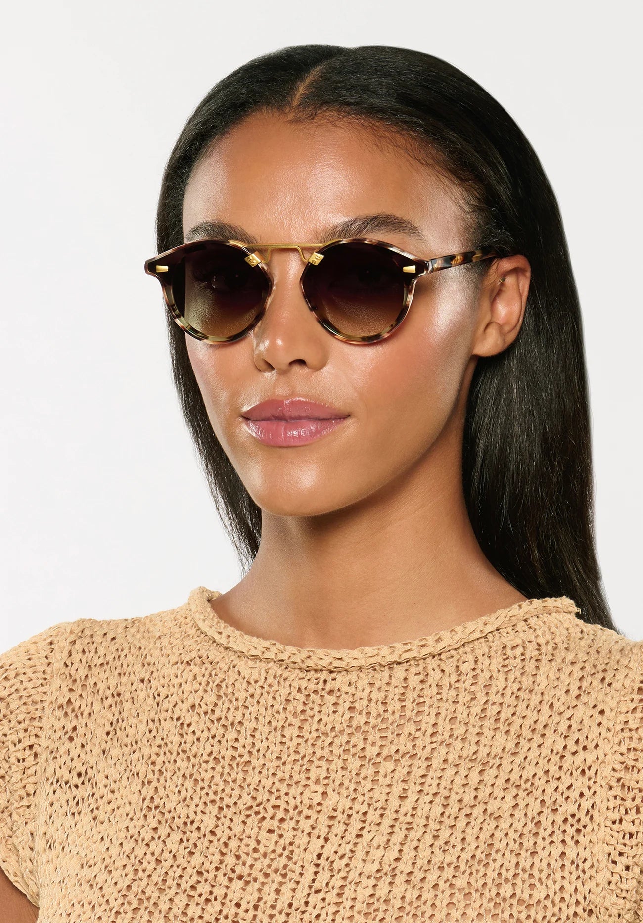 A woman with straight dark hair wears Krewe STL Nylon Sunglasses with flat nylon lenses and an oversized silhouette, paired with a beige knit top, facing forward against a plain light background.