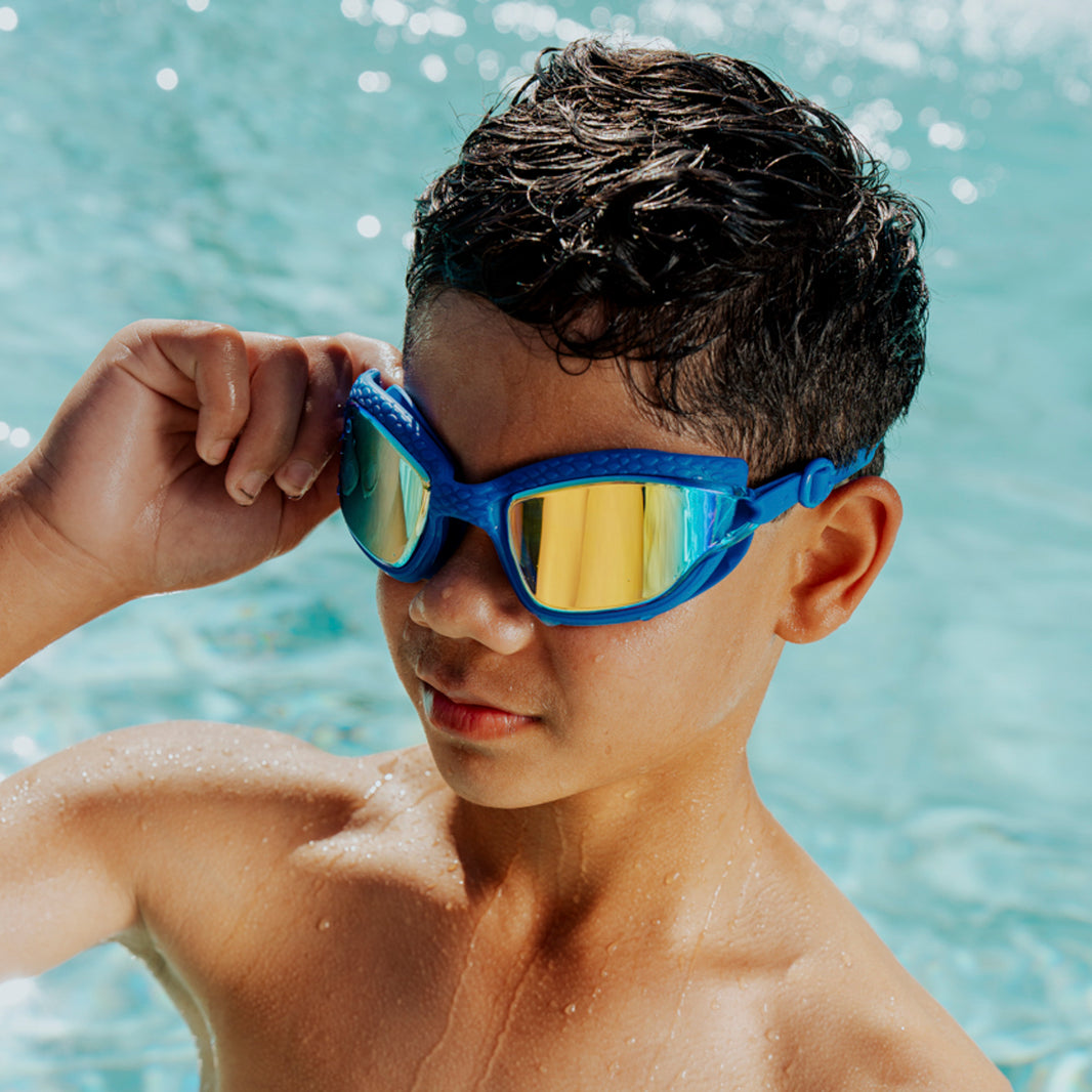 A boy wearing Bling2O Kids' Streamline Goggles in blue and yellow stands in a pool, with wet hair and water droplets on his skin.