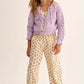 A young girl stands against a plain light background wearing the Cozmo Girls' Ivy Tricot Cardigan, featuring an openwork design, layered over a floral jumpsuit and paired with yellow sneakers.