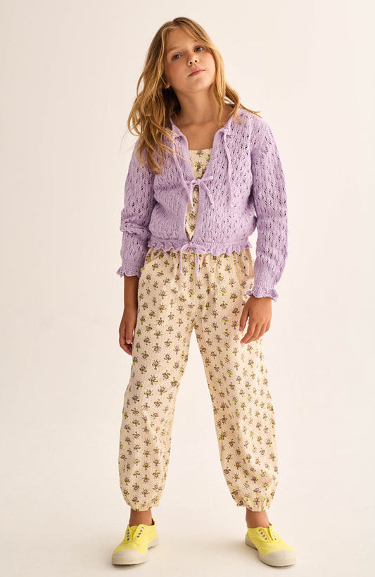 A young girl stands against a plain light background wearing the Cozmo Girls' Ivy Tricot Cardigan, featuring an openwork design, layered over a floral jumpsuit and paired with yellow sneakers.
