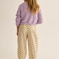 A young girl with long blond hair stands facing away against a plain light background, wearing a Cozmo Girls' Ivy Tricot Cardigan in lavender over cream floral-print pants and yellow shoes.