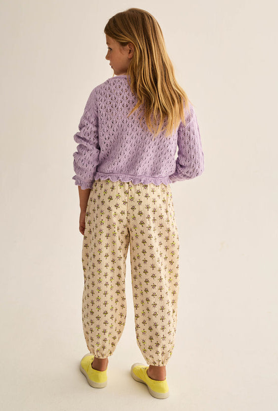A young girl with long blond hair stands facing away against a plain light background, wearing a Cozmo Girls' Ivy Tricot Cardigan in lavender over cream floral-print pants and yellow shoes.