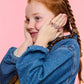 A young girl with braided red hair, wearing denim and the Super Smalls Heartfelt Sparkle Ring Set, holds her face with both hands against a pink background—perfect for kids' dress-up fun.