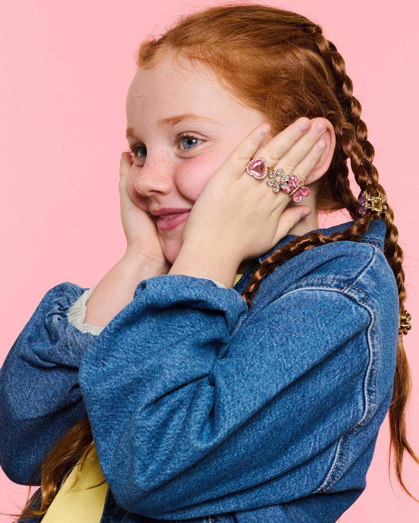 A young girl with braided red hair, wearing denim and the Super Smalls Heartfelt Sparkle Ring Set, holds her face with both hands against a pink background—perfect for kids' dress-up fun.