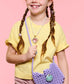 A young girl with red hair in braids, wearing a yellow shirt and pink pants, smiles while holding the Super Smalls Mermaid Dreams Handbag and wearing jewelry, standing against a pink background.