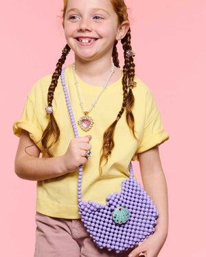 A young girl with red hair in braids, wearing a yellow shirt and pink pants, smiles while holding the Super Smalls Mermaid Dreams Handbag and wearing jewelry, standing against a pink background.