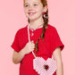 A smiling girl with braided hair holds the Super Smalls Heartfelt Sparkle Pearl Handbag, wearing a red shirt and red-and-white striped skirt against a pink background.