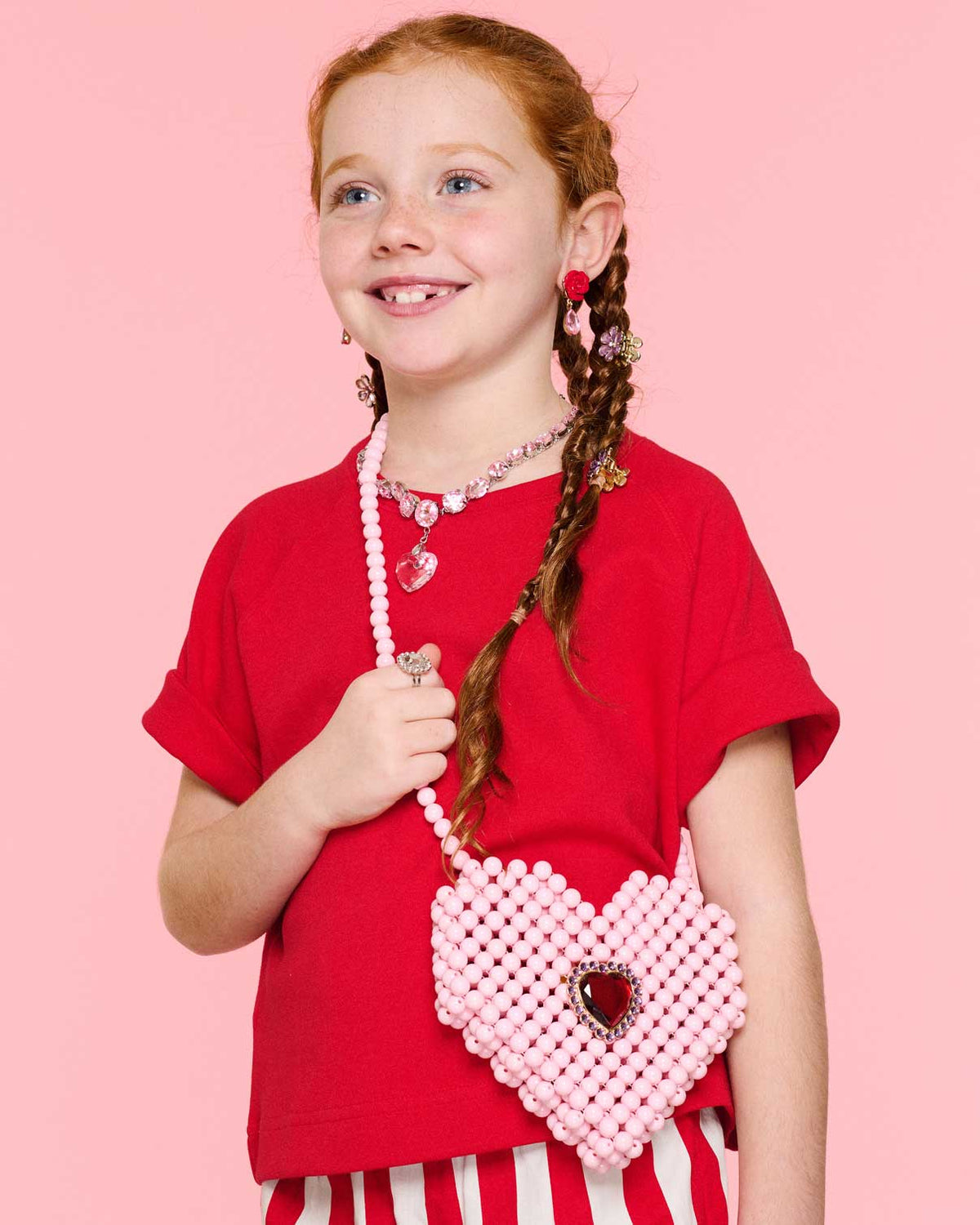 A smiling girl with braided hair holds the Super Smalls Heartfelt Sparkle Pearl Handbag, wearing a red shirt and red-and-white striped skirt against a pink background.