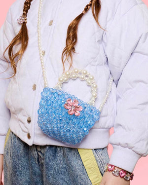 A person wears a light purple jacket and jeans, accessorized with the Super Smalls Opening Night Beaded Purse featuring pearl handles, a matching bracelet, and a crossbody strap, set against a pink background.