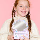 A smiling girl with braided hair holds the Super Smalls Mini Mermaid Vanity Gemify Kit, its mirror open, in front of a pink background decorated with sparkling gem stickers.