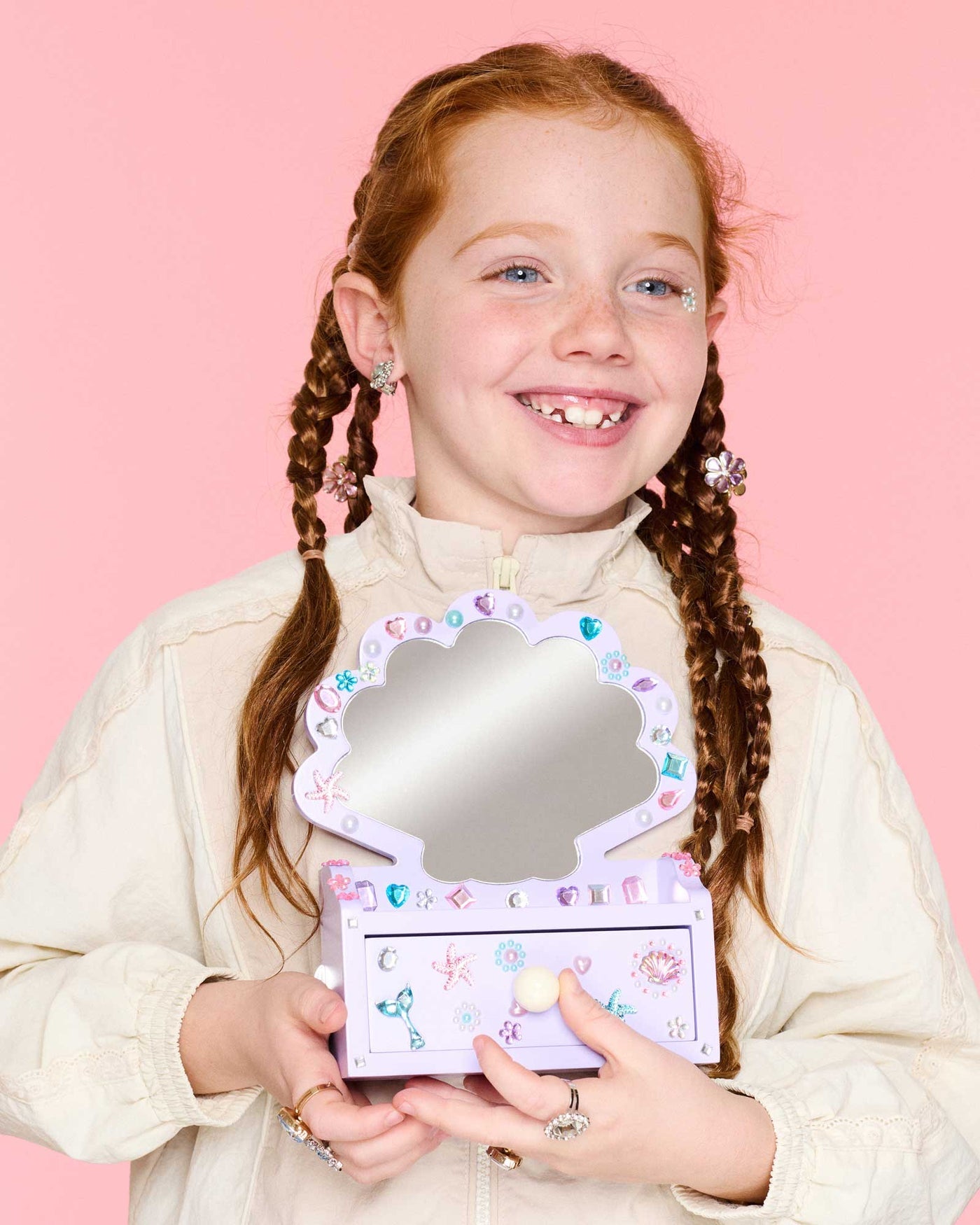 A smiling girl with braided hair holds the Super Smalls Mini Mermaid Vanity Gemify Kit, its mirror open, in front of a pink background decorated with sparkling gem stickers.