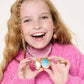 A smiling girl with long blonde hair in a pink sweater holds the Super Smalls Sparkle Shell Lipgloss Locket, revealing two stones inside. Her decorative nails, jewelry, and coconut-scented gloss add a playful touch to her look.
