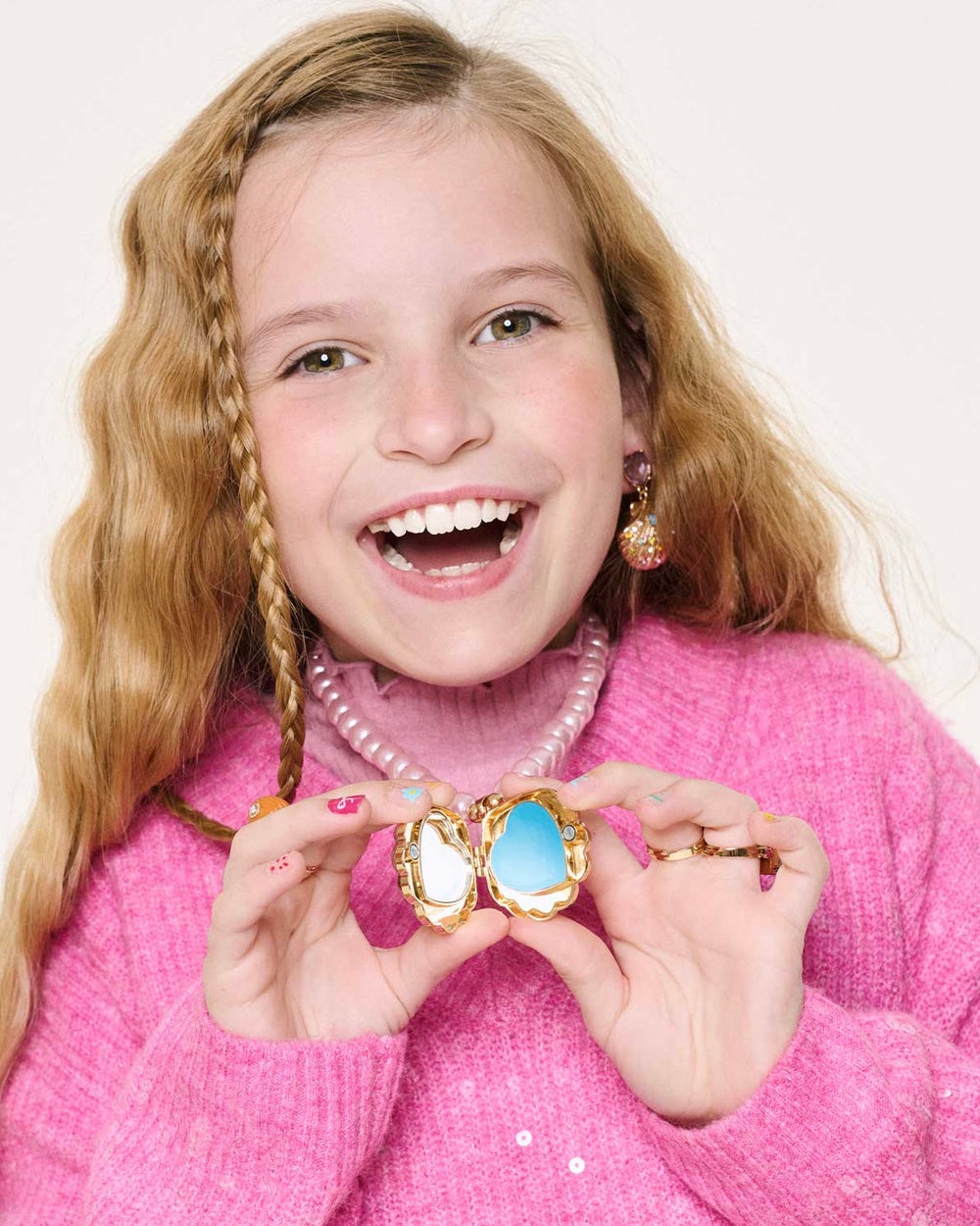 A smiling girl with long blonde hair in a pink sweater holds the Super Smalls Sparkle Shell Lipgloss Locket, revealing two stones inside. Her decorative nails, jewelry, and coconut-scented gloss add a playful touch to her look.
