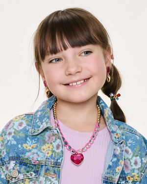 A young girl wearing a floral denim jacket, pink shirt, and vibrant accessories from the Super Smalls Perfectly Pink Mega Set—including playful clip-on earrings and a bold pink heart-shaped necklace—smiles and looks slightly to the side against a plain background.