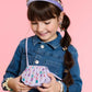 A young girl in a denim jacket smiles, holding the Super Smalls Mermaid Gemify Purse. She accessorizes with a bejeweled headband, gem stickers, earrings, and hair clips, standing against a pink background.