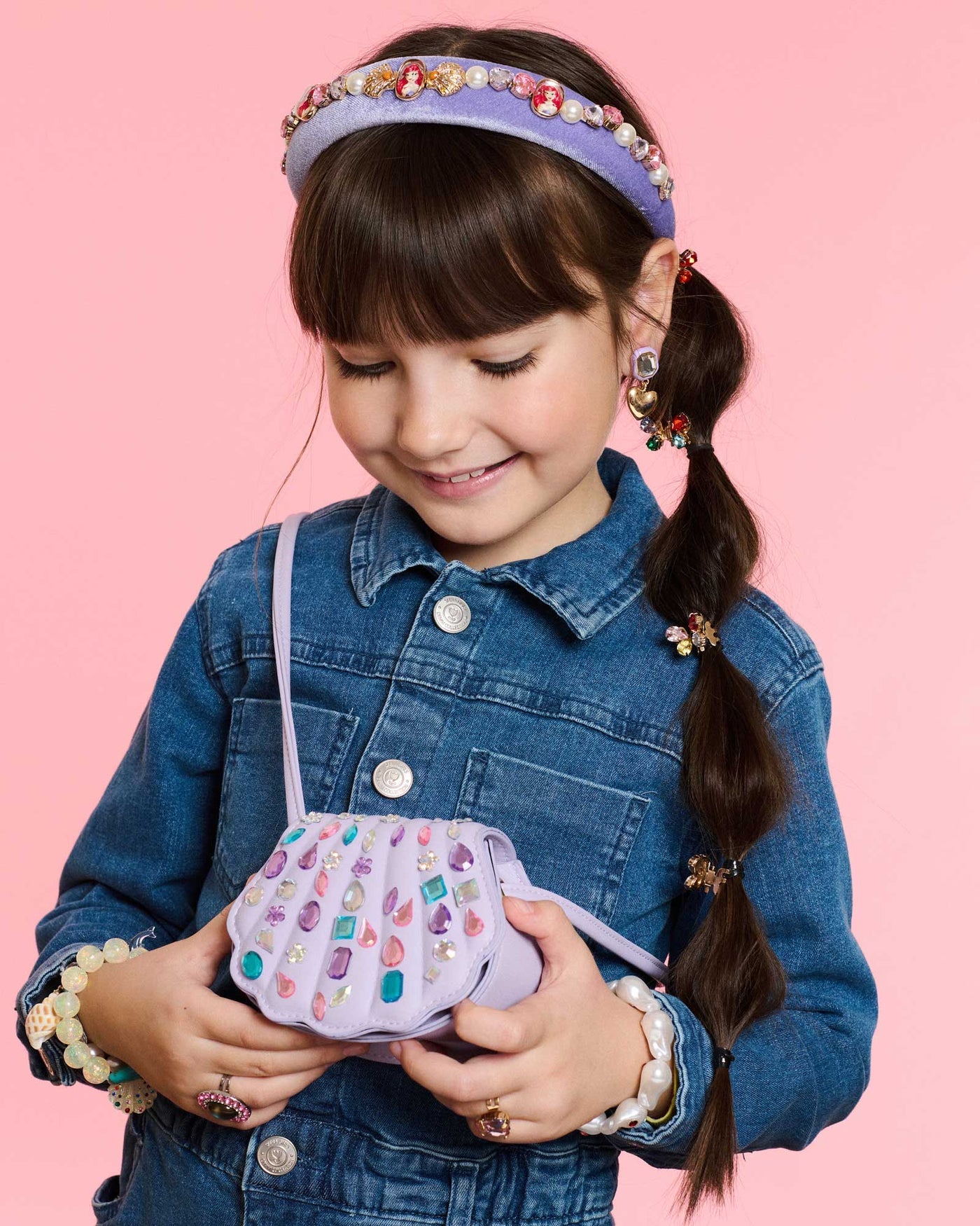 A young girl in a denim jacket smiles, holding the Super Smalls Mermaid Gemify Purse. She accessorizes with a bejeweled headband, gem stickers, earrings, and hair clips, standing against a pink background.