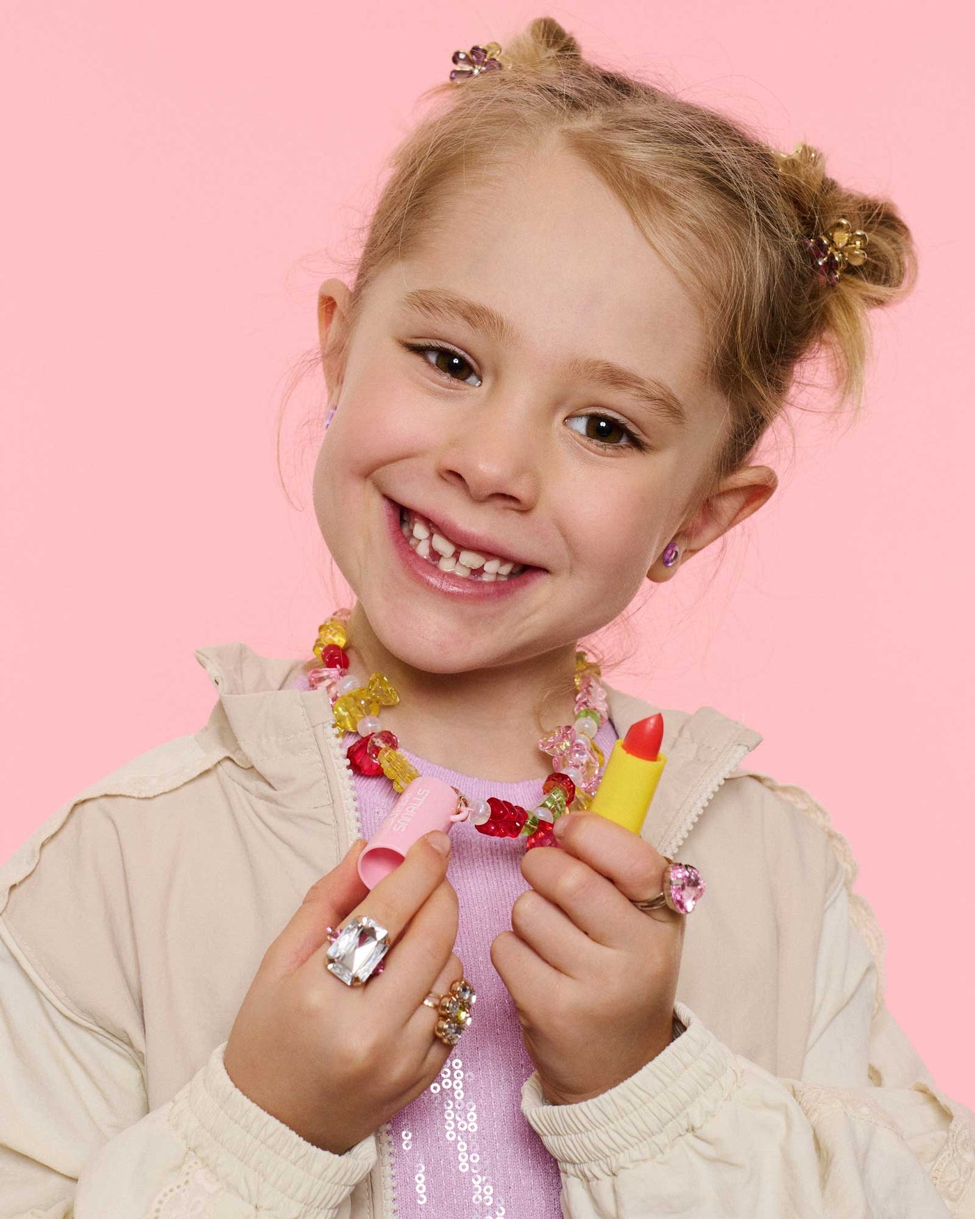 A smiling girl with a missing front tooth holds the Super Smalls Cutie Candy Lip Balm Bead Kit, wearing colorful jewelry she made herself, all set against a pink background.