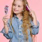 A young girl with long wavy hair in a denim jacket brushes her hair with a sparkly brush and mirror from the Super Smalls Unicorn Style & Shine Gemify Beauty Kit, set against a pink background.
