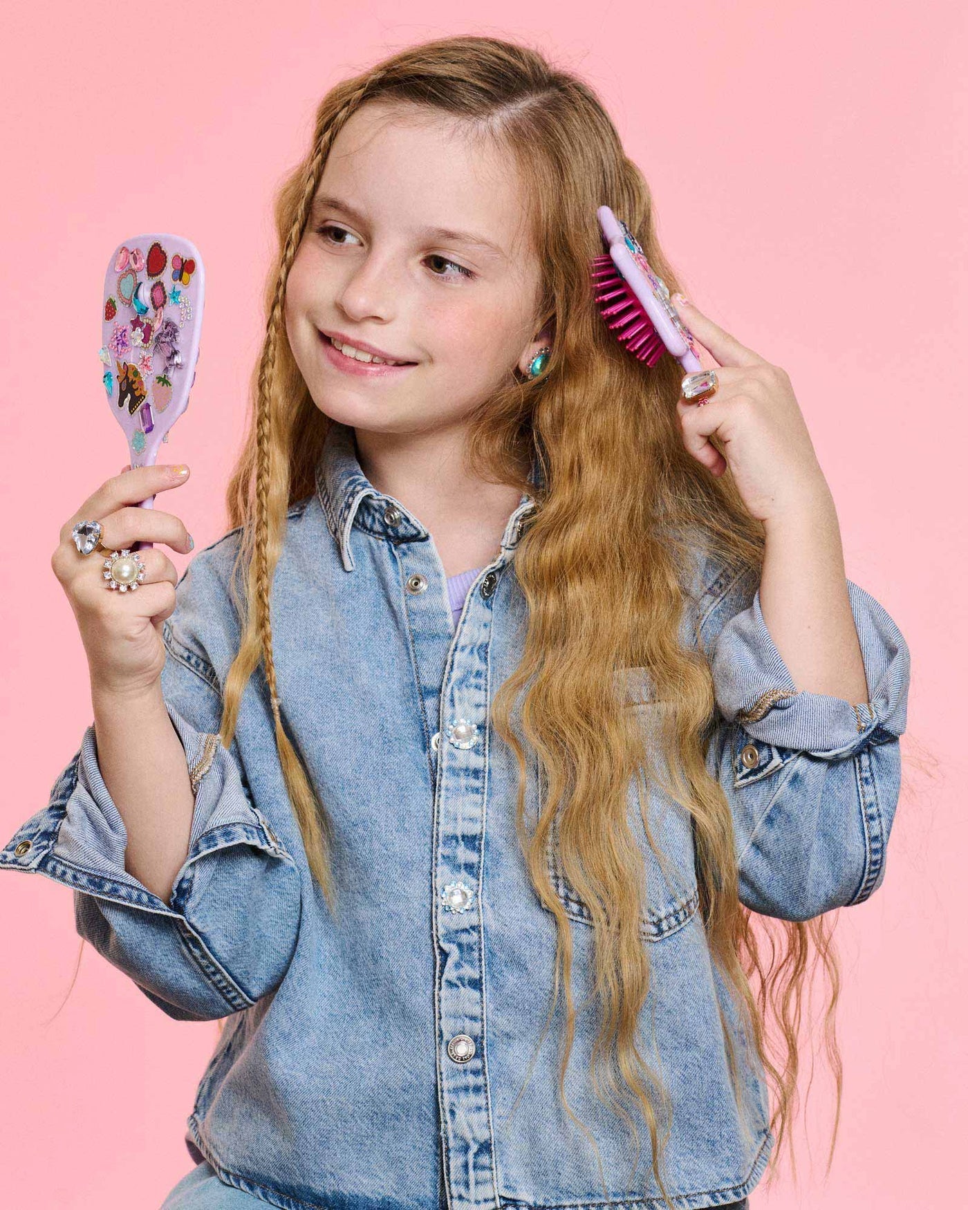 A young girl with long wavy hair in a denim jacket brushes her hair with a sparkly brush and mirror from the Super Smalls Unicorn Style & Shine Gemify Beauty Kit, set against a pink background.