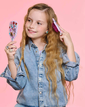A young girl with long wavy hair in a denim jacket brushes her hair with a sparkly brush and mirror from the Super Smalls Unicorn Style & Shine Gemify Beauty Kit, set against a pink background.