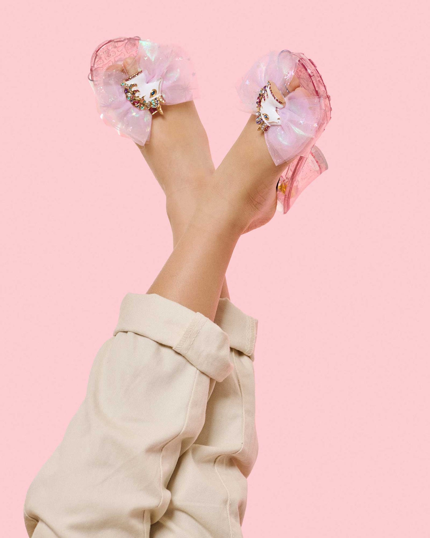A person wearing light-colored pants and Super Smalls Unicorn Wishes Playshoes with large sheer bows, glittery tulle, and unicorn decorations, shown on a pink background.