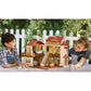 Two children sit at a table outdoors, playing with a Calico Critters Red Roof Country Home Gift Set and small animal figurines, with a white picket fence and green foliage in the background.