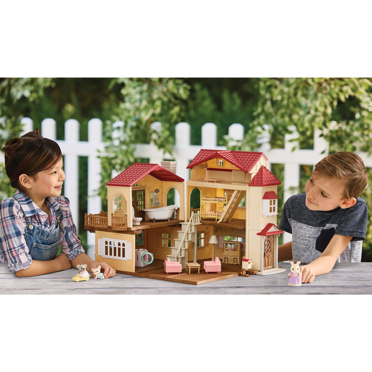 Two children sit at a table outdoors, playing with a Calico Critters Red Roof Country Home Gift Set and small animal figurines, with a white picket fence and green foliage in the background.