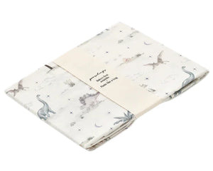 The Gooselings Dino Dreams Swaddle is a folded white 100% cotton muslin blanket, adorned with pastel dinosaurs and nature prints, wrapped in a cream paper band—ideal for adding a soothing touch to any nursery.