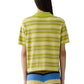 A person is seen from behind, wearing the Guest in Residence Short Sleeve Stripe Rugby in green and yellow over a cotton-cashmere tee, paired with a pleated color-block skirt in blue, yellow, and black.