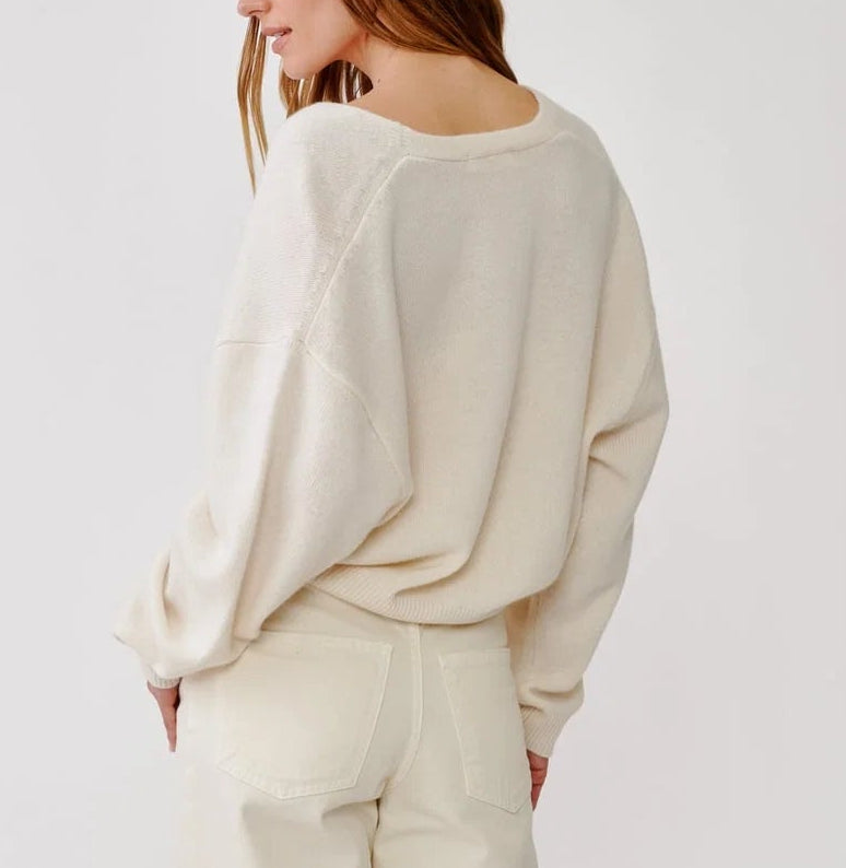 A person wearing the Sablyn Koda Wrap Cashmere Sweater—an oversized off-white wrap-front style with a deep V neckline—paired with cream pants stands facing away, highlighting the back and side of the outfit against a plain light background.