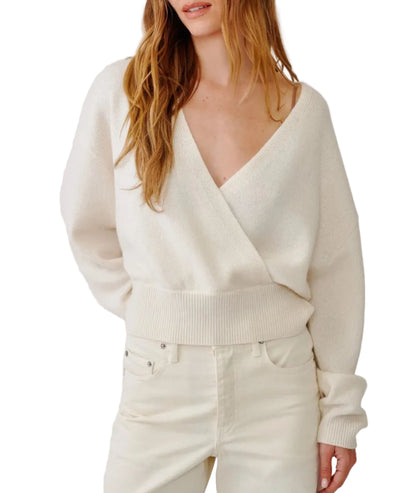 A woman wears the Sablyn Koda Wrap Cashmere Sweater—an elegant, cream-colored, long-sleeve wrap-front design with a deep V neckline—paired with high-waisted, off-white jeans. Her hair is loose and she stands against a white background.
