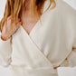 Wearing the Sablyn Koda Wrap Cashmere Sweater in cream, a woman with light pants stands slightly to the side, her hand resting on her shoulder and the sweater’s deep V neckline accentuating the wrap-front design.