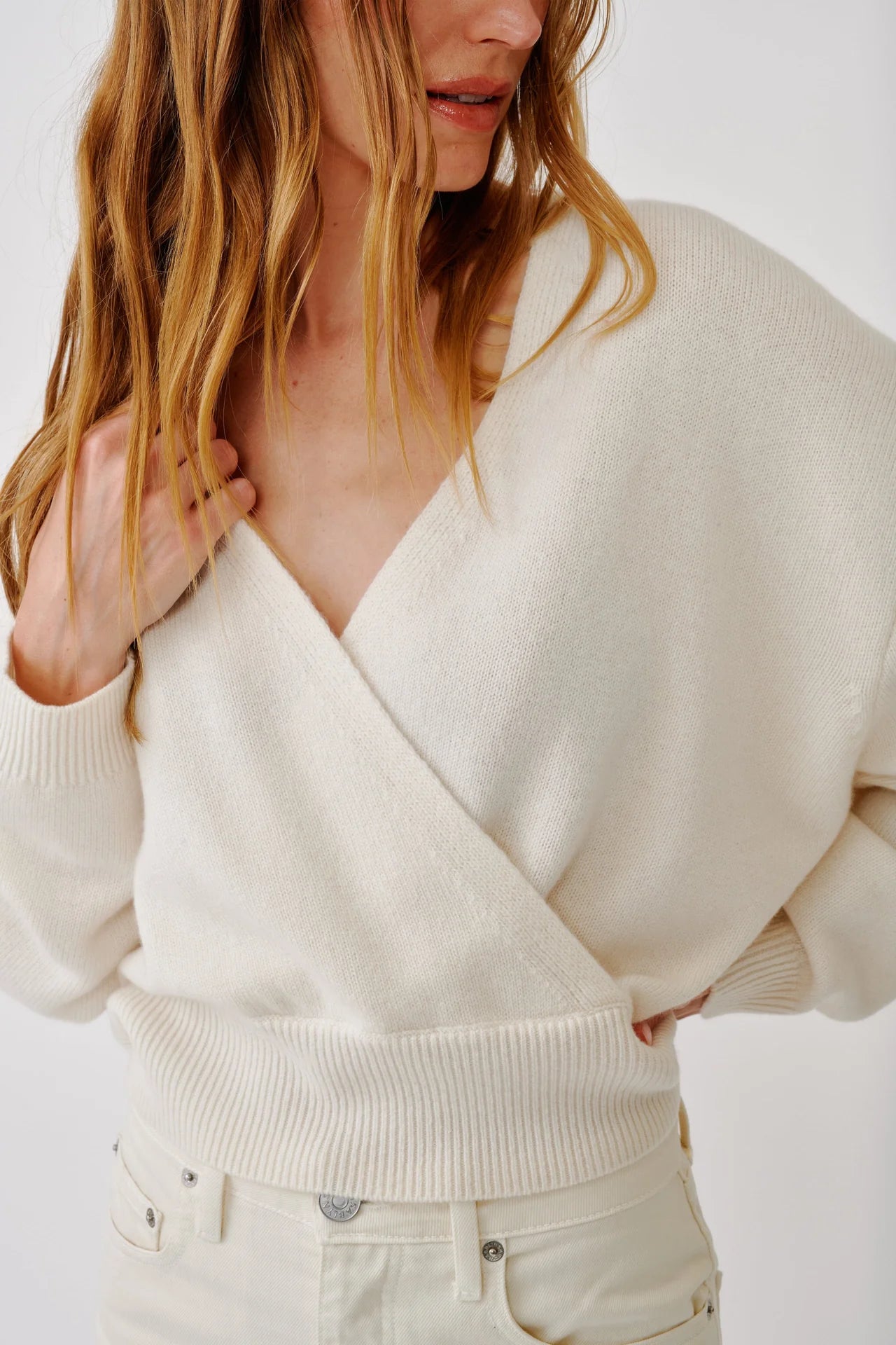 Wearing the Sablyn Koda Wrap Cashmere Sweater in cream, a woman with light pants stands slightly to the side, her hand resting on her shoulder and the sweater’s deep V neckline accentuating the wrap-front design.