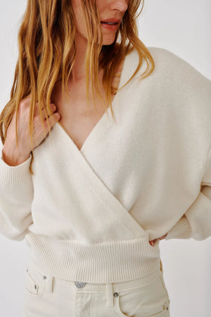 Wearing the Sablyn Koda Wrap Cashmere Sweater in cream, a woman with light pants stands slightly to the side, her hand resting on her shoulder and the sweater’s deep V neckline accentuating the wrap-front design.