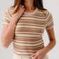 A woman wears the Sablyn Jude Short Sleeve Tee, featuring beige, brown, and cream multicolor stripes, paired with light pants against a plain background.