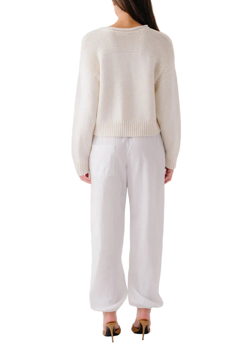 A woman, seen from the back against a white background, wears the Sablyn Sutton Slouchy Pullover in cream with ribbed detailing, paired with loose white pants and black high-heeled shoes.
