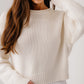 A woman wears the Sablyn Sutton Slouchy Pullover in cream with ribbed details, paired with white pants, standing against a plain light background.