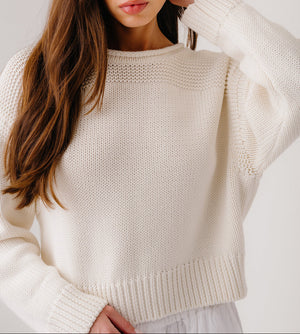 A woman wears the Sablyn Sutton Slouchy Pullover in cream with ribbed details, paired with white pants, standing against a plain light background.