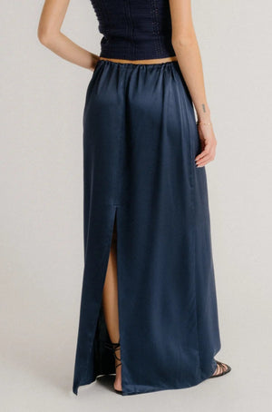 A person wearing the Sablyn Sol Drawstring Maxi Skirt in a relaxed fit with a back slit, paired with black strappy sandals and a sleeveless dark top, shown from the back.