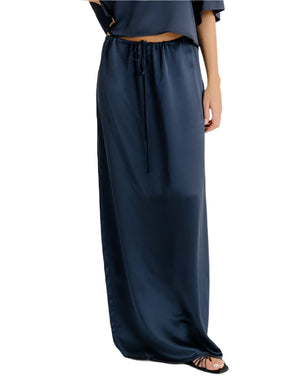 Someone wearing the Sablyn Sol Drawstring Maxi Skirt in navy blue silk, featuring an ankle-length cut and adjustable waistband, paired with black sandals.