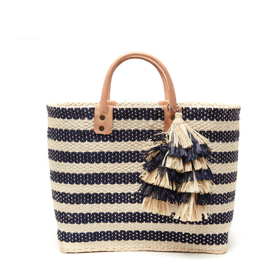 The Mar Y Sol Sahara Tote is a woven straw beach bag featuring navy and beige stripes, leather handles, and decorative straw tassels on one handle.