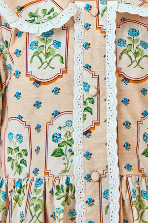 A close-up of the Girls' Sally Dress by Hunter Bell NYC highlights pastel botanicals, white lace trim, and fabric-covered buttons on a beige background—ideal for a mommy & me look.