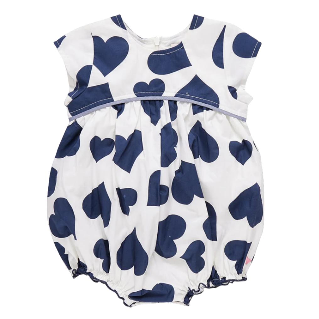 The Pink Chicken Baby Girls' Sana Bubble is a white romper adorned with large navy hearts, cap sleeves, and a gathered waist. This charming outfit also features delightful chambray details that add an extra touch of style.