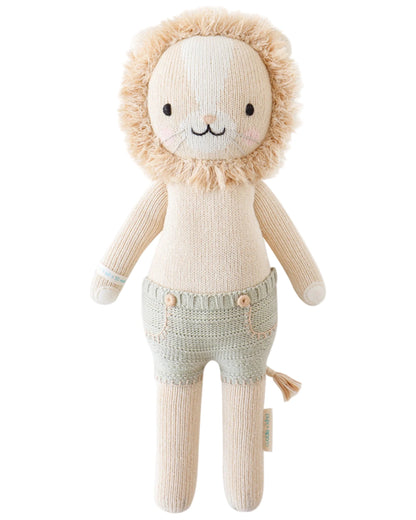 Sawyer the Lion - Regular 20" by Cuddle + Kind stands upright with a beige mane, light brown body, and green shorts against a white background—an ideal birthday gift that inspires kindness.