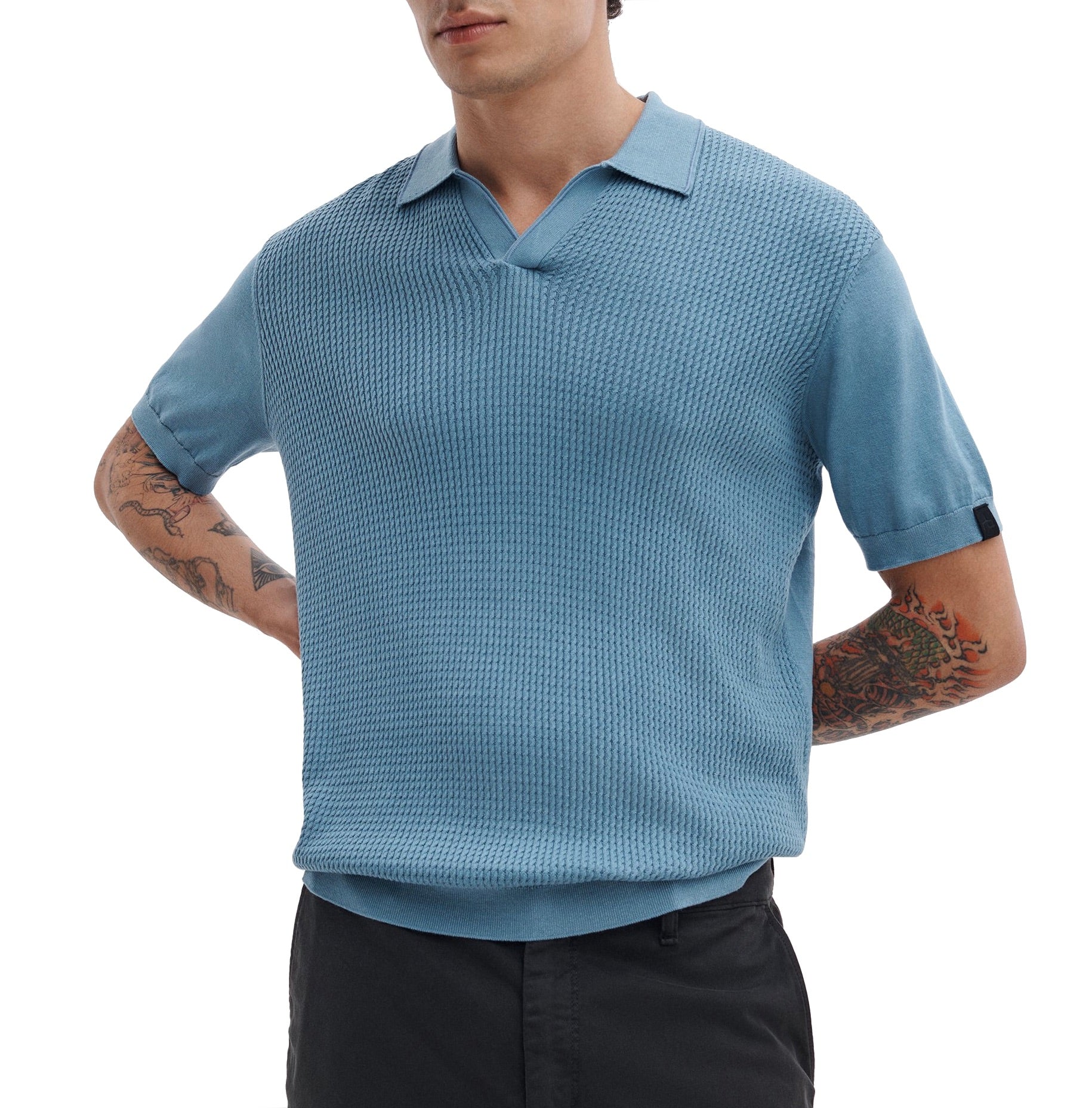 A man wearing a Rag & Bone Saxon Silk-Cotton Polo Shirt in light blue and black pants stands with his hands behind his back.