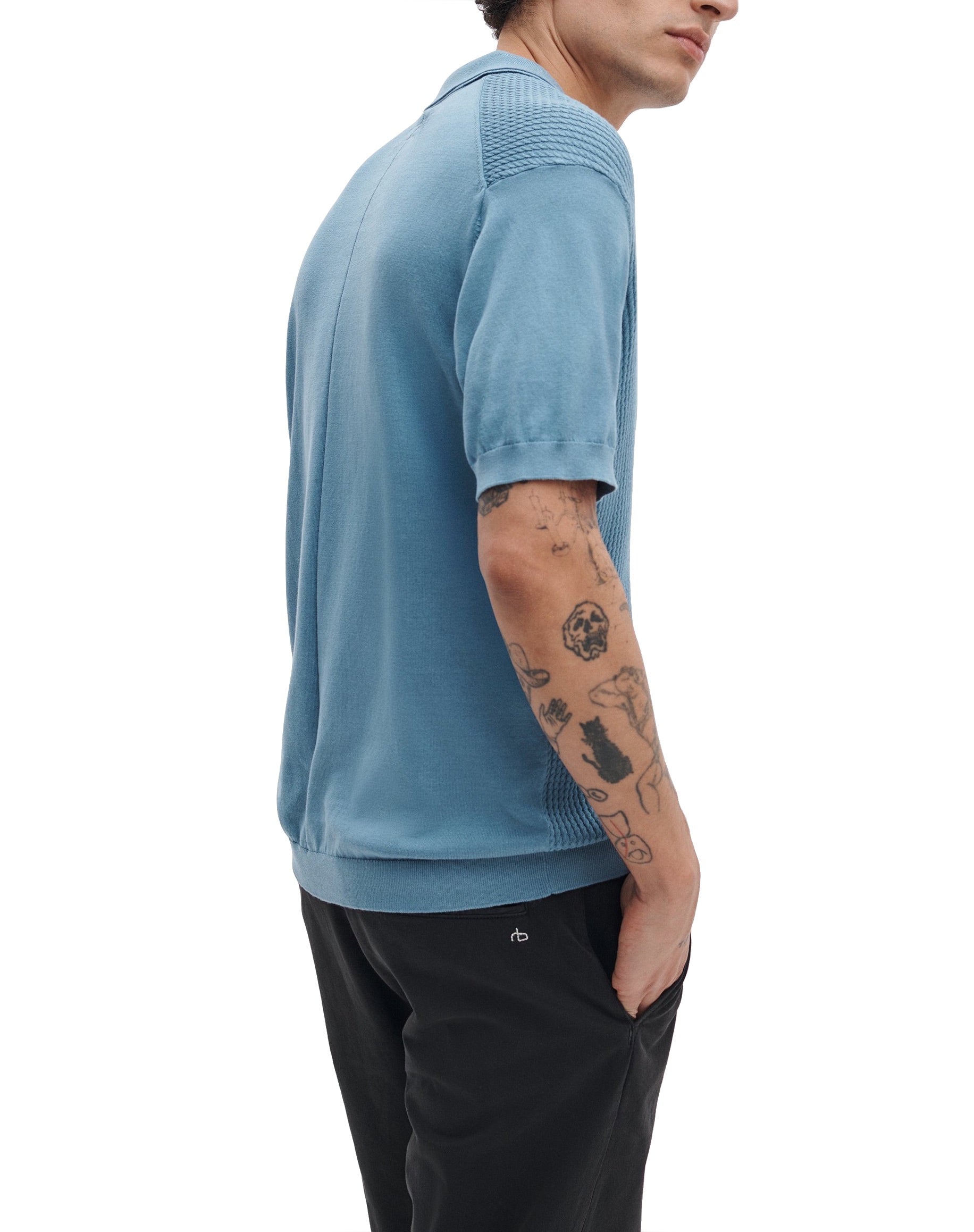 Wearing a Rag & Bone Saxon Silk-Cotton Polo Shirt and black pants, a person with several arm tattoos stands with one hand in their pocket.