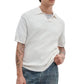 A person wearing the Rag & Bone Saxon Silk-Cotton Polo Shirt in white over a light gray T-shirt and blue jeans, with visible arm tattoos, stands against a plain white background.
