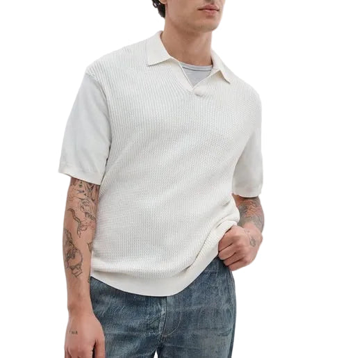 A person wearing the Rag & Bone Saxon Silk-Cotton Polo Shirt in white over a light gray T-shirt and blue jeans, with visible arm tattoos, stands against a plain white background.