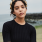 A person wearing a black shirt and the Gigi Burris Ava Band stands near a rocky beach, showcasing exquisite millinery craftsmanship.